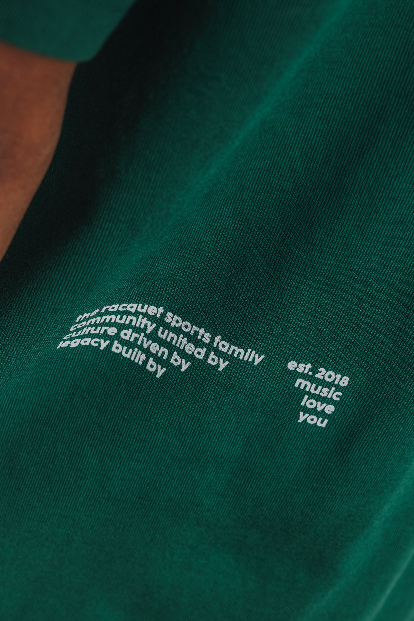 Close-up of small printed brand text at lower back hem of court green tee.