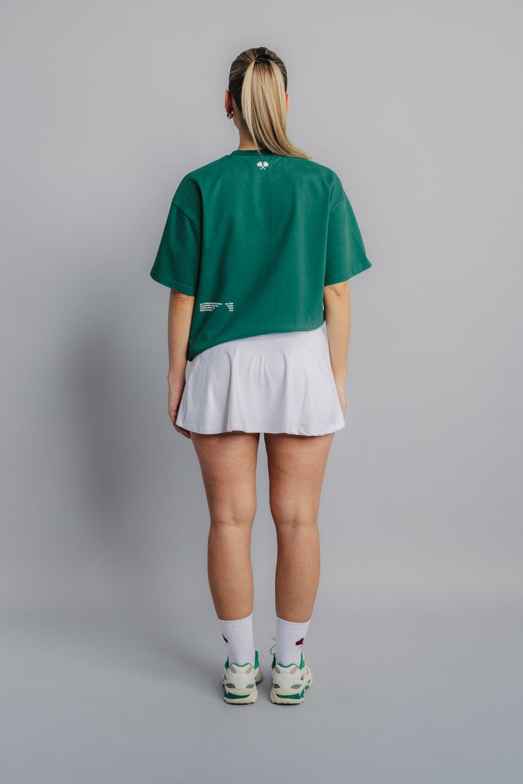 Female model showing back of court green tee with subtle heart racquet symbol near collar.