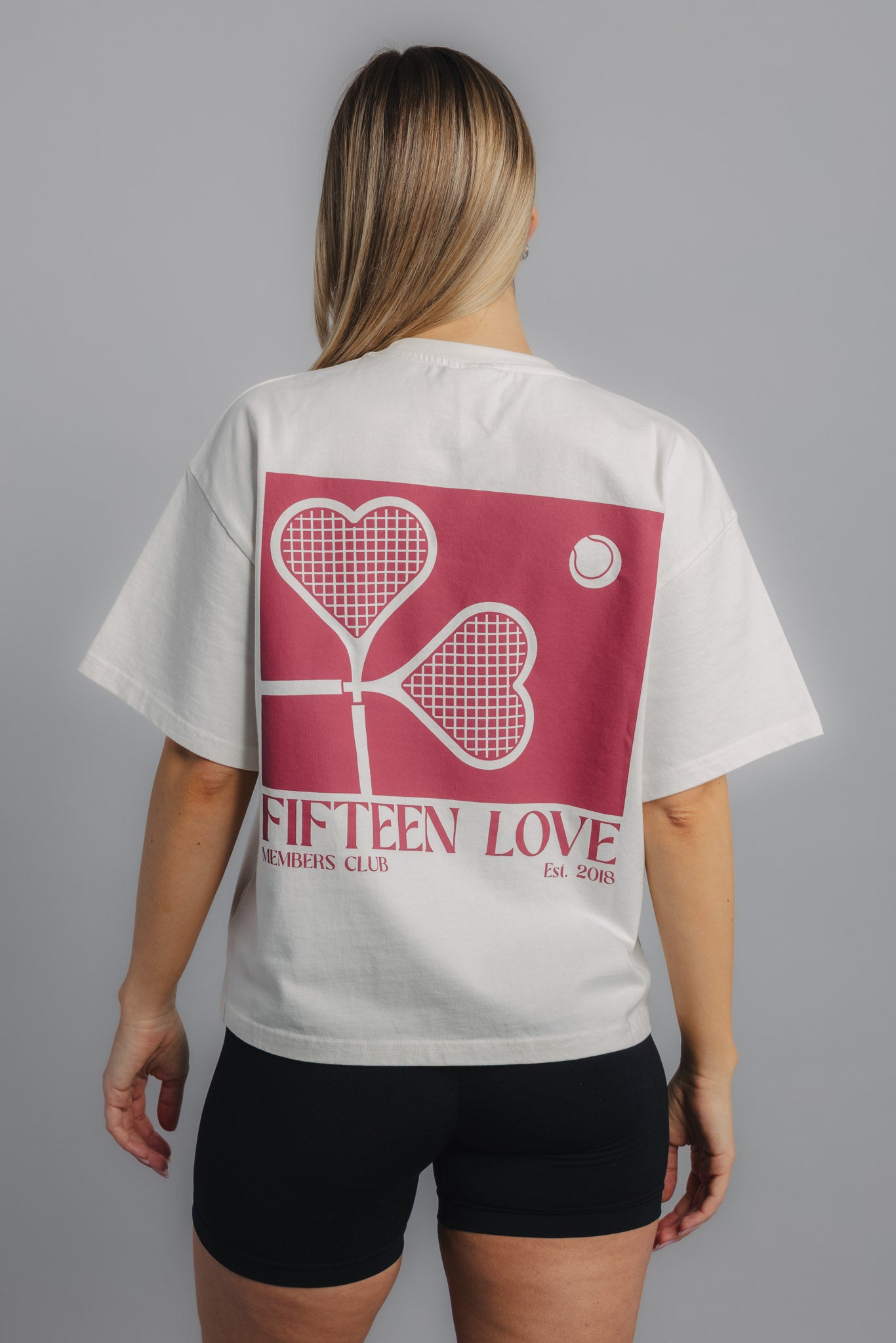 Female tennis model showing back of white tennis tee with bold heart racquet print.